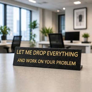 May include: A black rectangular desk sign with gold lettering that reads, "LET ME DROP EVERYTHING AND WORK ON YOUR PROBLEM." The sign is angled and features a textured surface, positioned on a flat surface in an office environment.