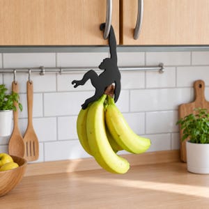 May include: A matte black metal monkey-shaped hook hanging from a kitchen cabinet handle, supporting a bunch of yellow bananas. The monkey silhouette features a curved tail for suspension and a hook-like hand to hold the fruit stem securely.