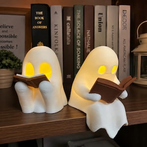 May include: Two white ghost-shaped lamps with glowing yellow eyes, each holding a small brown book. The ghost on the left sits upright, while the ghost on the right sits with its legs hanging over the edge of a wooden surface.
