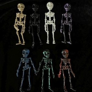 May include: A collection of seven plastic skeleton figurines in various colors, including off-white, black, purple, blue, green, and red. The skeletons are posed upright with articulated limbs, perfect for Halloween decorations or themed crafts.