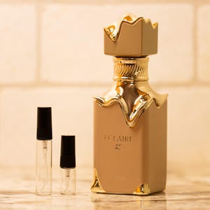 May include: A perfume bottle with a gold crown-shaped cap and gold accents. The bottle is a light brown color with the word "ECLAIRE" printed on the front. Two small sample bottles with black caps are in front of the perfume bottle.