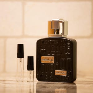 Puede incluir: Un frasco de perfume negro con una tapa plateada y dorada, acompañado de dos pequeños frascos de muestra con atomizador. El frasco de perfume tiene texto en relieve y una etiqueta dorada que dice "Ramz Lattafa".