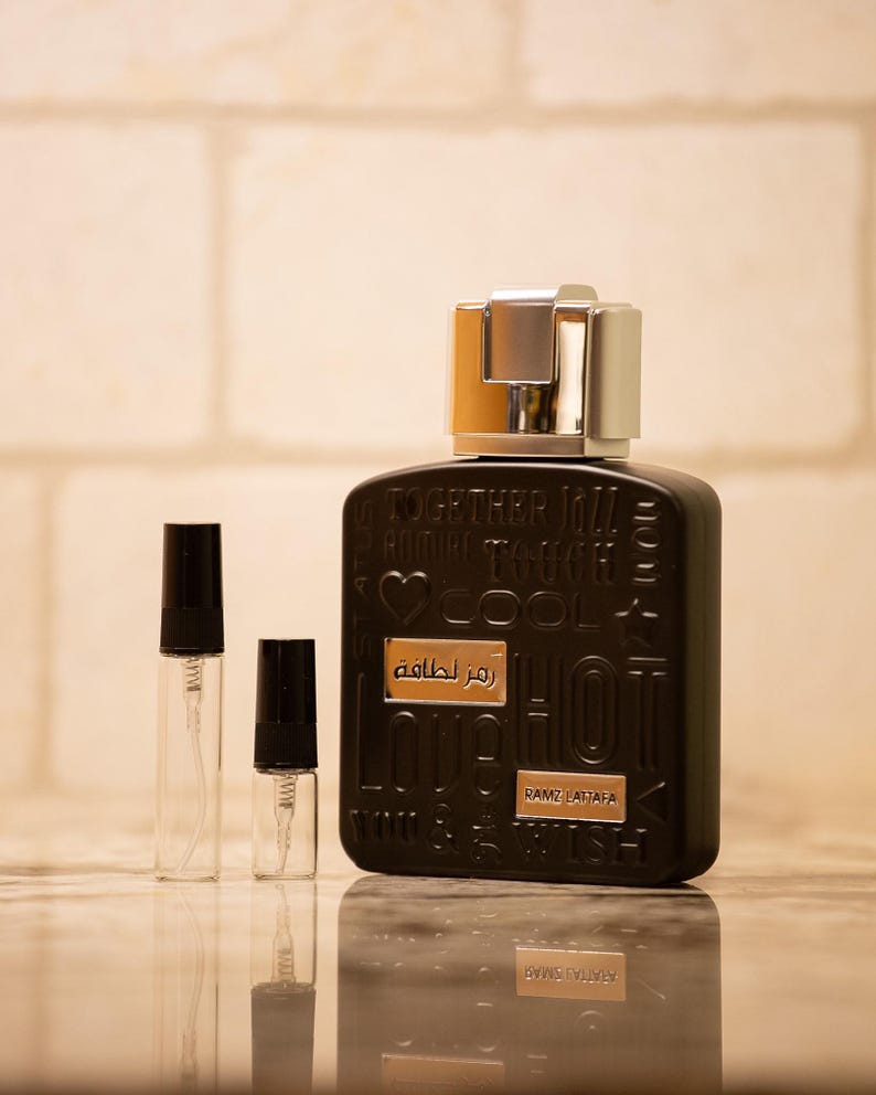 May include: A black perfume bottle with a silver and gold cap, accompanied by two small sample spray bottles. The perfume bottle has embossed text and a gold label that reads "Ramz Lattafa".
