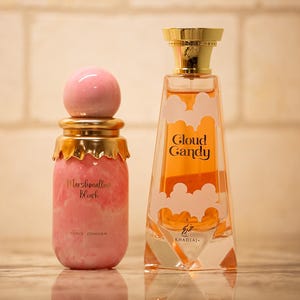 Peut inclure: Deux flacons de parfum. L'un est rose avec un bouchon doré et l'inscription "Marshmallow Blush". L'autre est un flacon transparent en forme de trapèze avec l'inscription "Cloud Candy" et un bouchon doré.