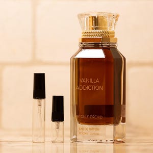May include: A perfume bottle with the text "Vanilla Addiction" and two smaller spray bottles. The large bottle is brown with a gold cap and a clear, faceted base. The smaller bottles have black caps.