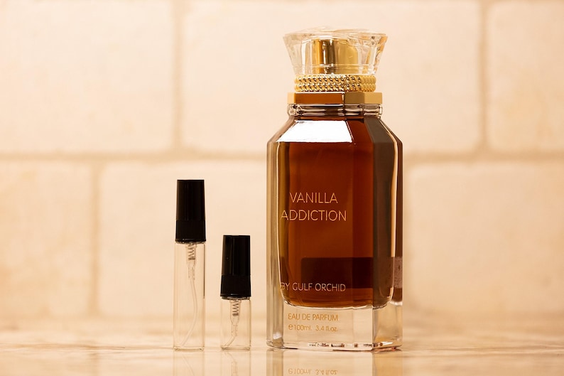 May include: A perfume bottle with the text "Vanilla Addiction" and two smaller spray bottles. The large bottle is brown with a gold cap and a clear, faceted base. The smaller bottles have black caps.