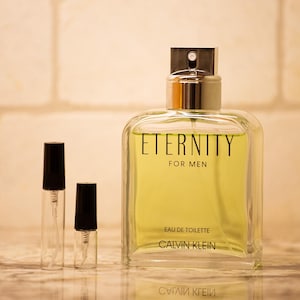 Puede incluir: Una botella de vidrio transparente de eau de toilette "Eternity for Men" de Calvin Klein, con tapa plateada y líquido amarillo claro. Dos botellas de spray más pequeñas y vacías con tapas negras al frente.