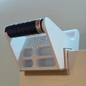 May include: A white plastic door-mounted exercise handle featuring a black textured grip wrap. The device has a rectangular frame with four internal openings and the letters BSI embossed on the top surface. It is secured over the top edge of a door.