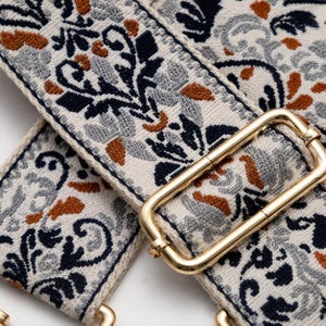 May include: A wide woven bag strap featuring a cream-colored base with a detailed floral pattern in navy blue, light gray, and burnt orange. The strap includes a polished gold-tone metal rectangular slider buckle for length adjustment.