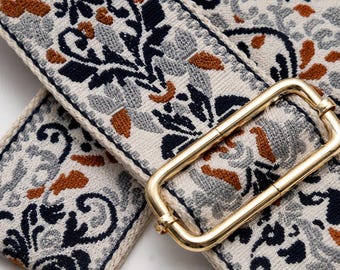 May include: A wide woven bag strap featuring a cream-colored base with a detailed floral pattern in navy blue, light gray, and burnt orange. The strap includes a polished gold-tone metal rectangular slider buckle for length adjustment.