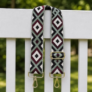 May include: A wide, adjustable bag strap featuring a repeating geometric diamond pattern in shades of burgundy, sage green, black, and white. The strap includes gold-toned metal hardware, including a sliding buckle and two swivel lobster clasps.