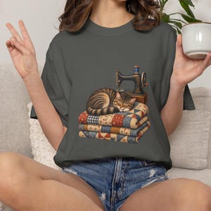 May include: A charcoal gray t-shirt featuring a graphic print of a brown tabby cat curled up asleep on a stack of three folded, multi-colored patchwork quilts. Behind the cat sits a vintage-style sewing machine with a blue thread spool.