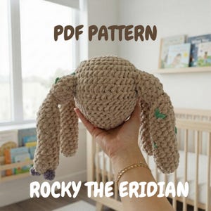 Può includere: Un giocattolo di peluche beige all'uncinetto con il testo "PDF PATTERN" e "ROCKY THE ERIDIAN". Il giocattolo ha un corpo rotondo e quattro arti allungati. Gli arti hanno accenti verdi e viola. Il giocattolo è tenuto in una mano.