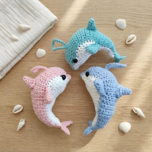 Puede incluir: Tres juguetes de delfines de ganchillo en tonos rosa, azul claro y azul, cada uno con vientres blancos y ojos negros. Los juguetes están dispuestos sobre una superficie de madera con conchas marinas y un paño blanco.