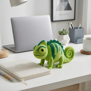 May include: A crocheted chameleon toy in shades of green and yellow, with a curled tail. It sits on a white desk alongside a notebook, a laptop, and a pen. The chameleon has a smiling face.