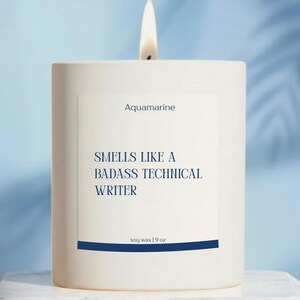 May include: A lit white candle with the text "Aquamarine" and "Smells Like a Badass Technical Writer" on a white label. The candle is in a white square container and is labeled "soy wax | 9 oz".