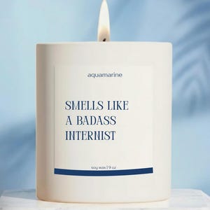 May include: A white rectangular candle with a lit wick. The label reads "aquamarine" and "Smells Like a Badass Internist." The candle is soy wax, 9 oz. A blue stripe is at the bottom of the label. The background is a light blue.
