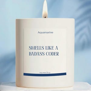 May include: A lit white candle with the text "Aquamarine" and "Smells Like a Badass Coder" in navy blue. The candle is soy wax and weighs 9 oz. The candle sits on a white marble surface against a light blue background.