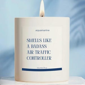 May include: A white square candle with a lit wick. The label reads "aquamarine" and "Smells Like A Badass Air Traffic Controller." The candle is soy wax, 9 oz.