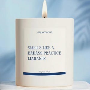 May include: A white candle with a lit wick. The label reads "aquamarine" and "Smells Like a Badass Practice Manager." It is soy wax and weighs 9 oz. The candle is on a white marble surface.