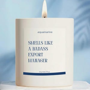 May include: A lit white candle with the text "Smells Like A Badass Export Manager" on a white label. The candle is labeled "aquamarine" and is made of soy wax, 9 oz.