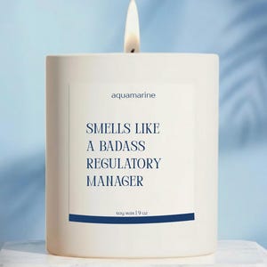 May include: A lit white candle with the text "aquamarine" and "Smells Like A Badass Regulatory Manager" on a white label. The candle is soy wax and weighs 9 oz.