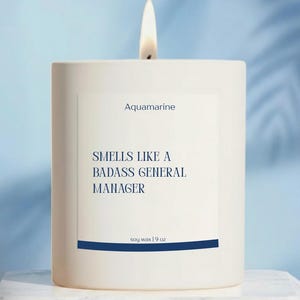 May include: A lit white candle with the text "Aquamarine" and "Smells Like a Badass General Manager." The candle is in a white square container with a blue stripe and the text "soy wax | 9 oz."