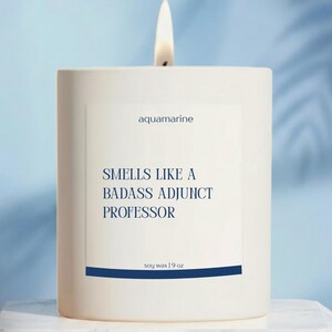 May include: A white candle with a lit wick. The label reads "aquamarine" and "Smells Like a Badass Adjunct Professor." The candle is soy wax, 9 oz. The background is a light blue.