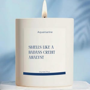 May include: A white square candle with a lit wick. The label reads "Aquamarine" and "Smells Like a Badass Credit Analyst." The candle is soy wax, 9 oz. The background is a light blue with a marble surface.
