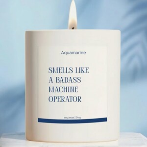 May include: A lit white candle with the text "Aquamarine" and "Smells Like A Badass Machine Operator" on a label. The candle is soy wax, 9 oz.