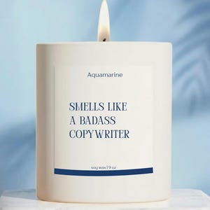 May include: A white square candle with a lit wick. The label reads "Aquamarine" and "Smells Like a Badass Copywriter." The candle is soy wax, 9 oz. The background is a light blue with a marble surface.