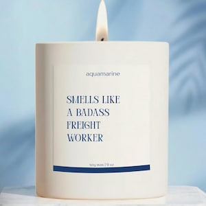 May include: A lit white candle with the text "aquamarine" and "Smells Like A Badass Freight Worker" in navy blue. The candle is soy wax, 9 oz.