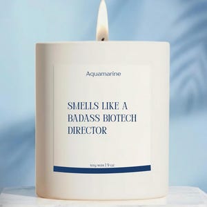 May include: A lit white candle with a label that reads "Aquamarine" and "Smells Like a Badass Biotech Director." The candle is made of soy wax and is 9 oz. The background is a light blue.
