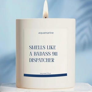 May include: A lit white candle with the text "aquamarine" and "Smells Like A Badass 911 Dispatcher." The candle is soy wax and weighs 9 oz. The background is a light blue.