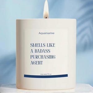 May include: A lit white candle with a label that reads "Aquamarine" and "Smells Like A Badass Purchasing Agent." The candle is in a white square container and is made of soy wax, 9 oz.