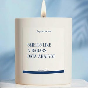 May include: A white square candle with a lit wick. The label reads "Aquamarine" and "Smells Like a Badass Data Analyst." The candle is soy wax and weighs 9 oz. The background is a light blue.