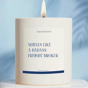 May include: A white square candle with a lit wick. The label reads "aquamarine" and "Smells Like a Badass Freight Broker." The candle is soy wax, 9 oz.