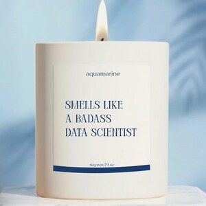 May include: A white soy wax candle with a lit wick. The label reads "aquamarine" and "Smells Like a Badass Data Scientist." The candle is 9 oz.