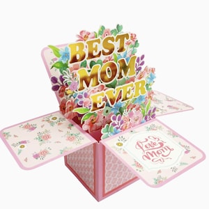 Könnte beinhalten: Eine rosa Pop-up-Muttertagskarte in Boxform mit den Worten "BEST MOM EVER" in goldfarbenen Buchstaben, umgeben von bunten Blumen. Die Karte hat florale Muster und die Worte "Love Mom".