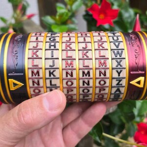 May include: A cylindrical puzzle with rotating lettered rings. The device is black, gold, and burgundy with a series of letters on each ring. The ends have gold triangles. The puzzle is held in a hand against a background of green leaves and red flowers.