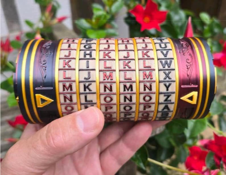 May include: A cylindrical puzzle with rotating lettered rings. The device is black, gold, and burgundy with a series of letters on each ring. The ends have gold triangles. The puzzle is held in a hand against a background of green leaves and red flowers.
