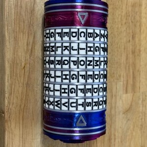 May include: A cylindrical puzzle with rotating lettered rings. The top and bottom sections are blue, with a magenta band in the middle. The rings are white with black letters. A triangle is visible on the magenta band.
