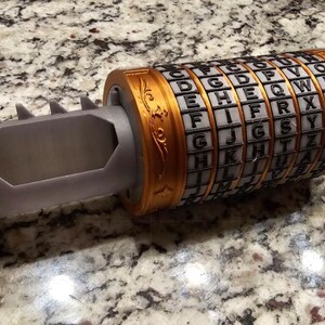 May include: A cylindrical puzzle with rotating rings, featuring an alphabet cipher. The device is primarily grey with gold-coloured accents on the ends and rings. The rings display letters, and the device has a hollow interior.