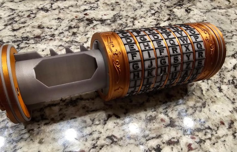 May include: A cylindrical puzzle with rotating rings, featuring an alphabet cipher. The device is primarily grey with gold-coloured accents on the ends and rings. The rings display letters, and the device has a hollow interior.