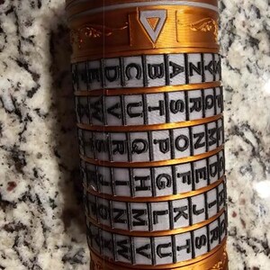 May include: A cylindrical puzzle in gold and grey. The puzzle has rotating bands with the alphabet. Triangular shapes decorate the top and bottom. The puzzle is designed to be solved, with the letters used to unlock it.