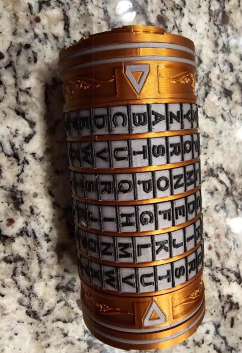 May include: A cylindrical puzzle in gold and grey. The puzzle has rotating bands with the alphabet. Triangular shapes decorate the top and bottom. The puzzle is designed to be solved, with the letters used to unlock it.