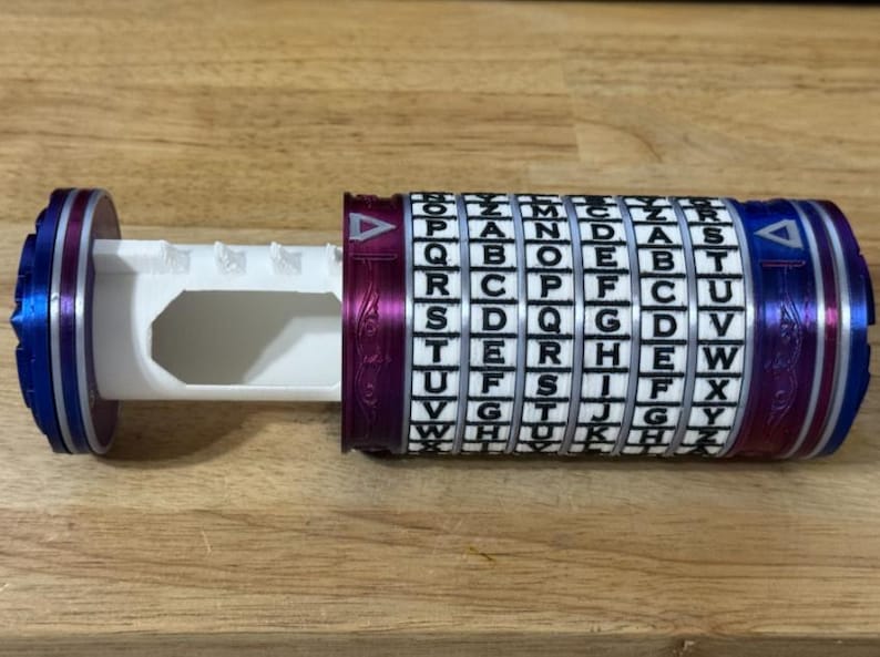 May include: A cylindrical puzzle box with a rotating alphabet cipher. The box is primarily white, with blue and purple accents. The cipher has letters in a grid, and the box is partially open, revealing an interior compartment.