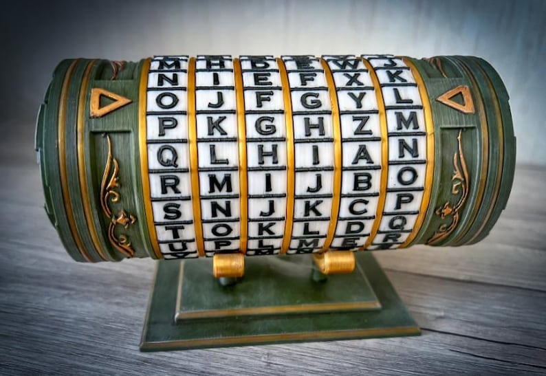 May include: A green and gold cipher cylinder with rotating lettered rings. The device has a rectangular base and decorative gold accents. The letters are black on white blocks. The overall design suggests a puzzle or secret code.