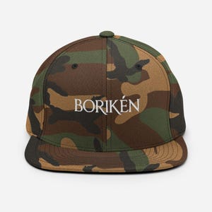 May include: A camouflage print baseball cap featuring a flat brim and a structured crown. The word BORIKÉN is embroidered in white capital letters across the front centre of the cap. The pattern consists of brown, dark green, and black shapes.