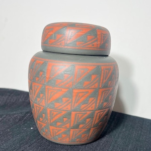 May include: A matte gray ceramic jar with a matching lid, featuring a repeating geometric pattern in burnt orange. The design consists of interconnected triangles and angular lines covering the entire surface of the vessel and its lid.